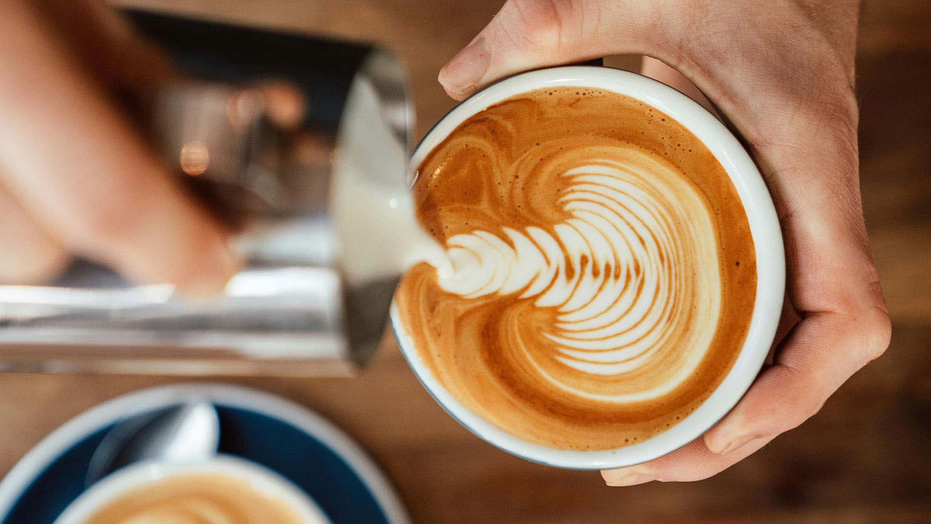 Close-up of a hand pouring steamed milk into an espresso drink, expertly crafting a feathered latte art design in a white ceramic cup