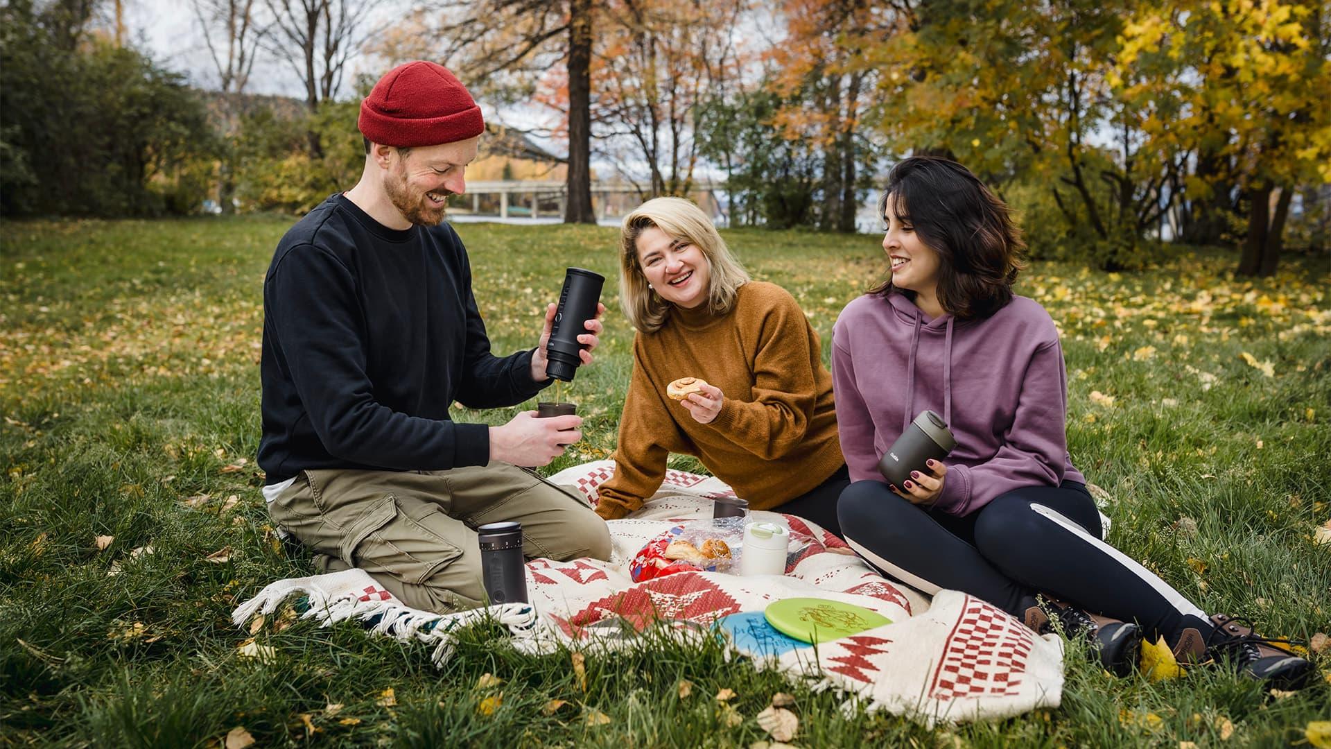 Group picnic in park, holding black portable coffee makers, enjoying autumn outdoors.