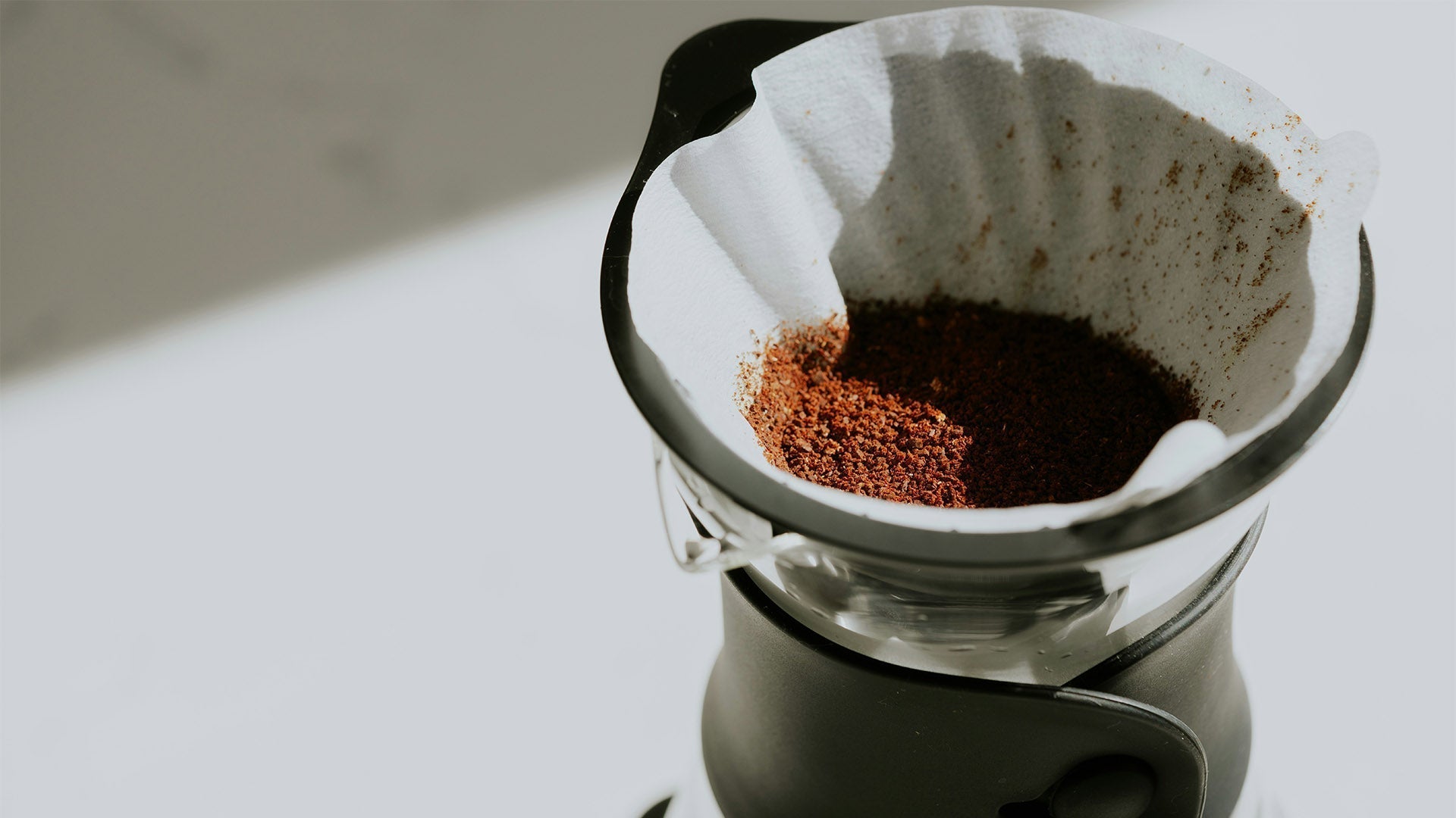 Coarse coffee grounds rest in a paper filter inside a pour-over brewer. Bright light casts a sharp diagonal shadow across the white surface