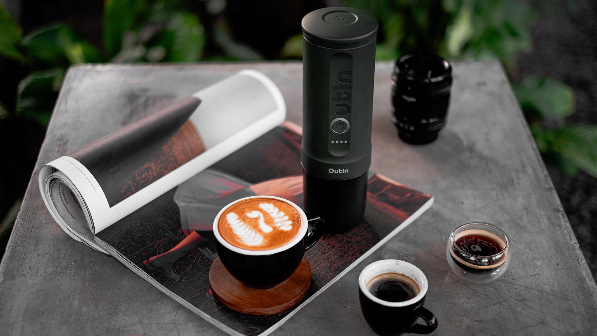 Outin portable espresso maker with logo next to a latte with latte art on a coffee table