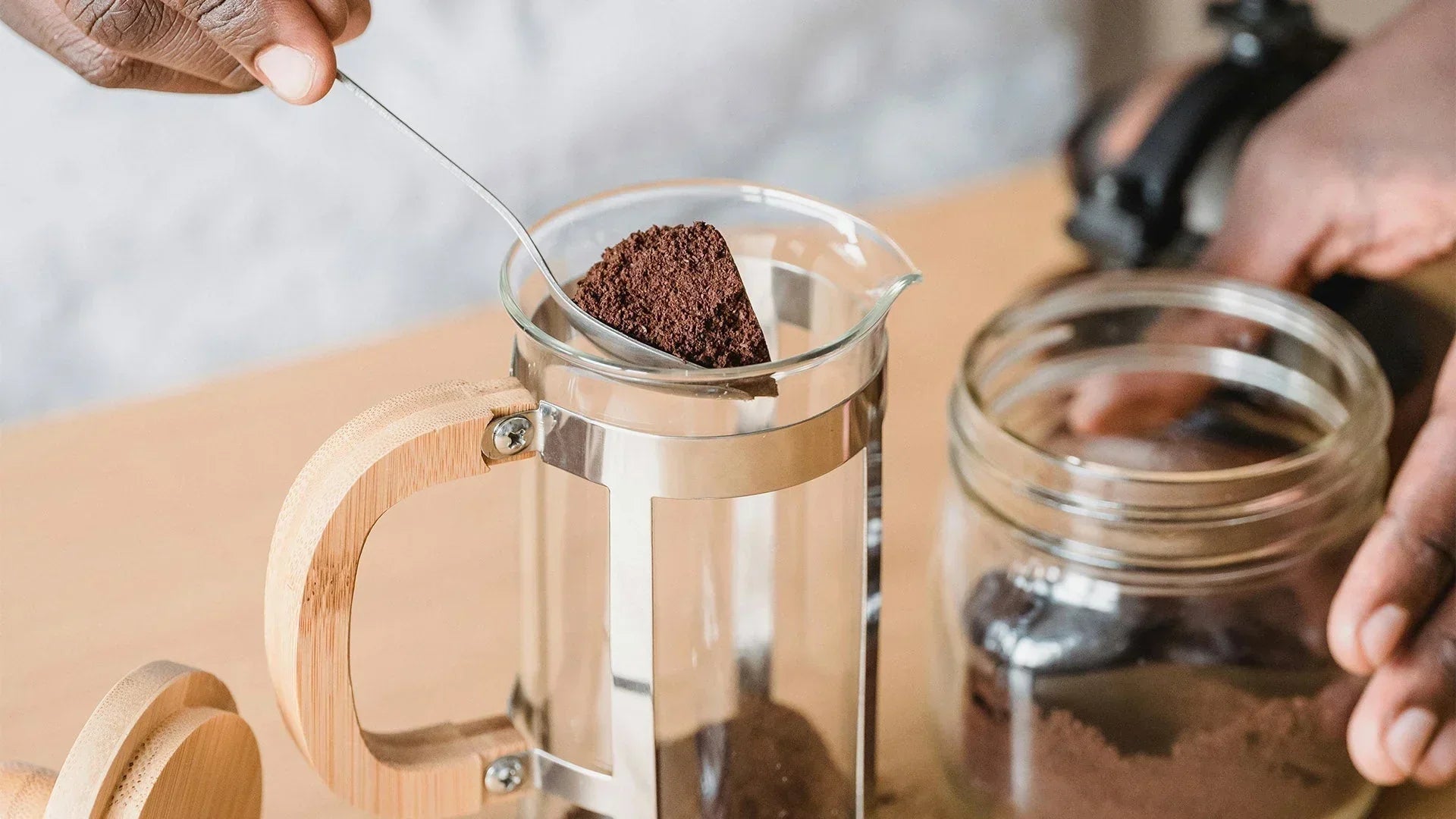 A person is spooning coarse coffee grounds from a jar into a glass French press with a wooden handle, preparing to brew