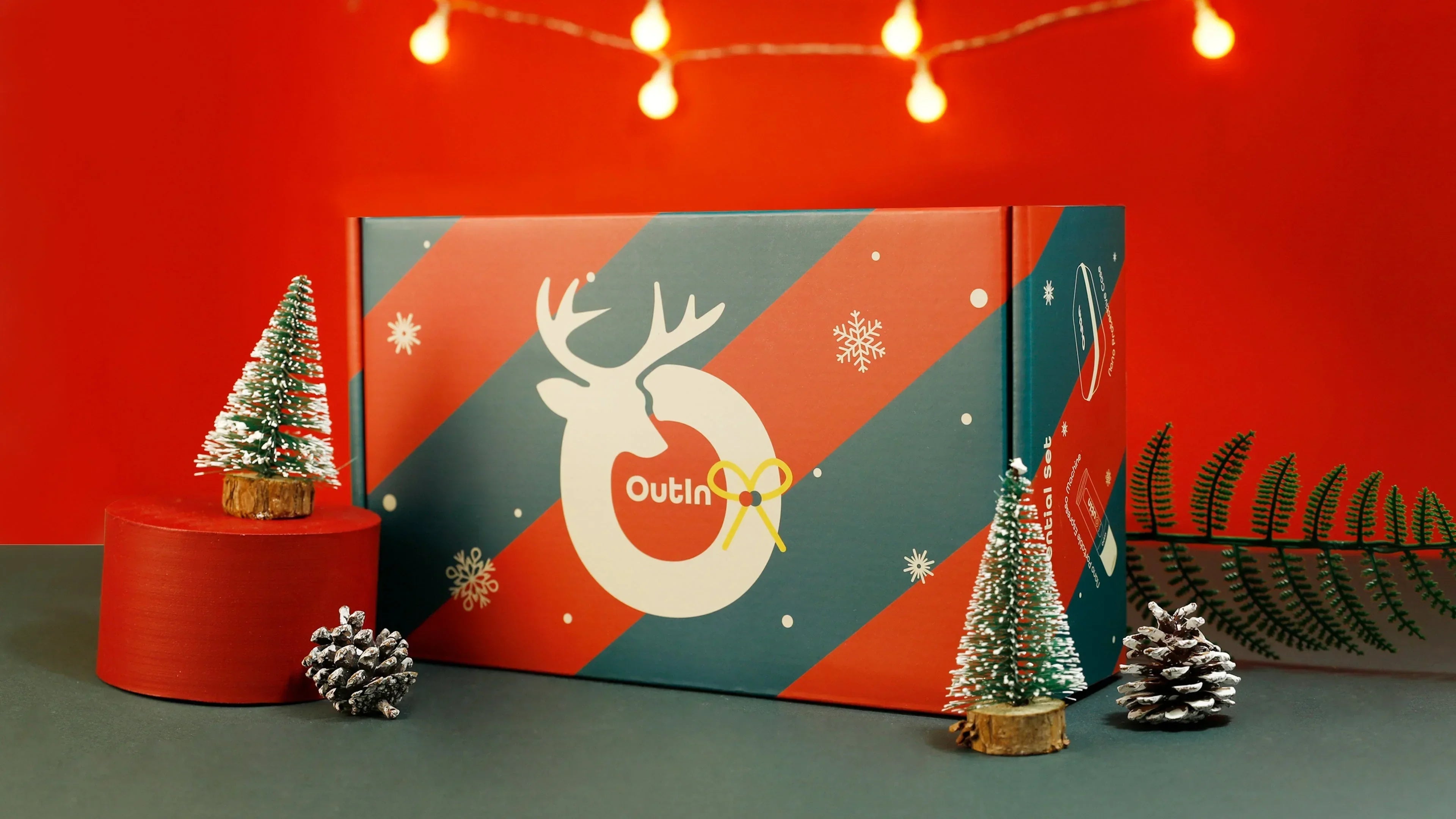 OutIn Nano black portable espresso machine with Christmas gift box, coffee capsules, mini Christmas tree and fairy lights on red backdrop