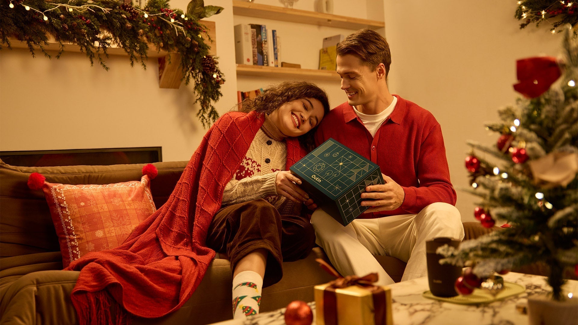 A cozy couple is sitting on a couch in a Christmas-decorated room, happily opening a dark green gift box labeled "Outin"