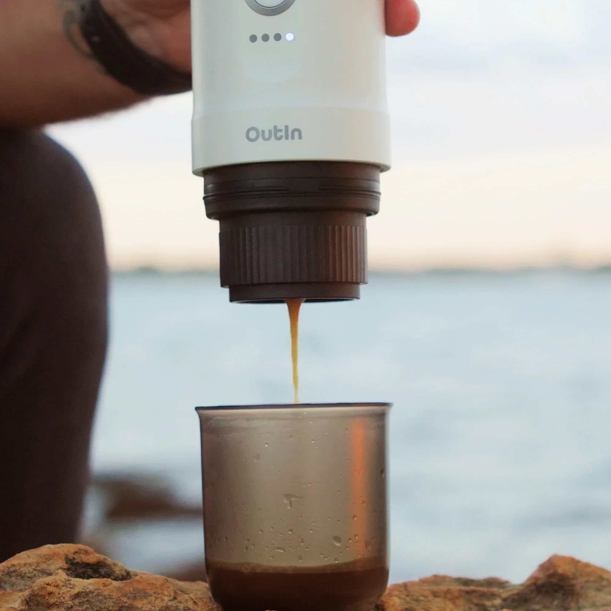 Pearl white Nano portable espresso machine brewing rich espresso with crema into a cup outdoors on rocks.