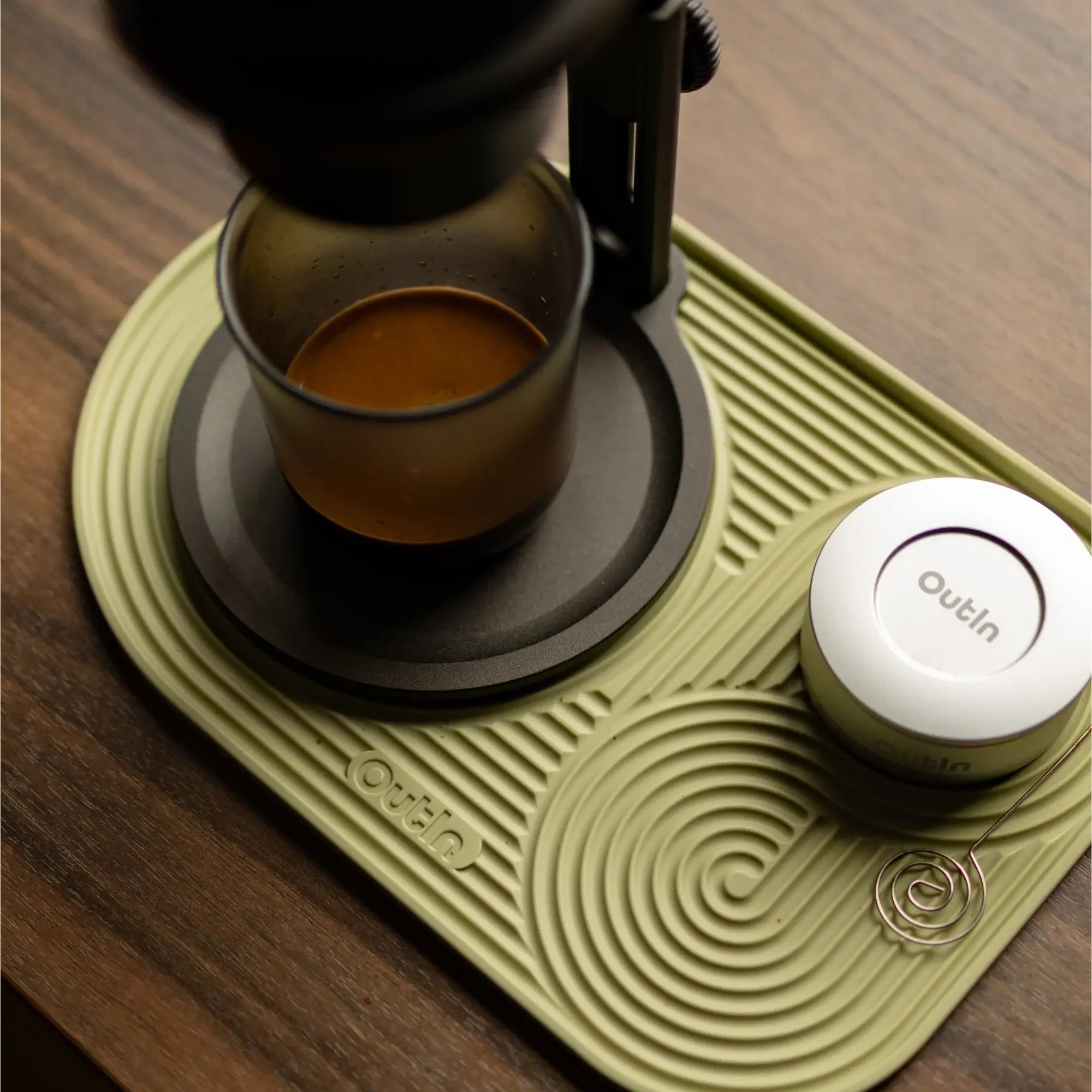 OutIn sage green silicone coffee mat with espresso glass, dosing cup, and whisk, showcasing product context on a wooden desk.