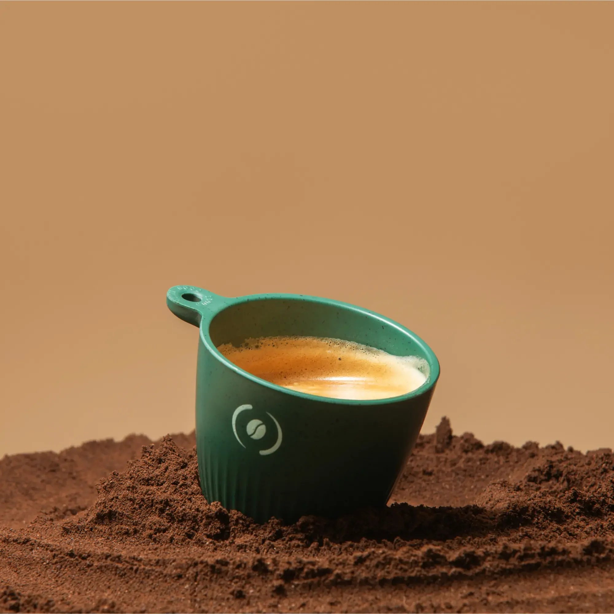 OutIn Rebrew dark green espresso cup filled with espresso, with coffee bean logo, on coffee grounds.