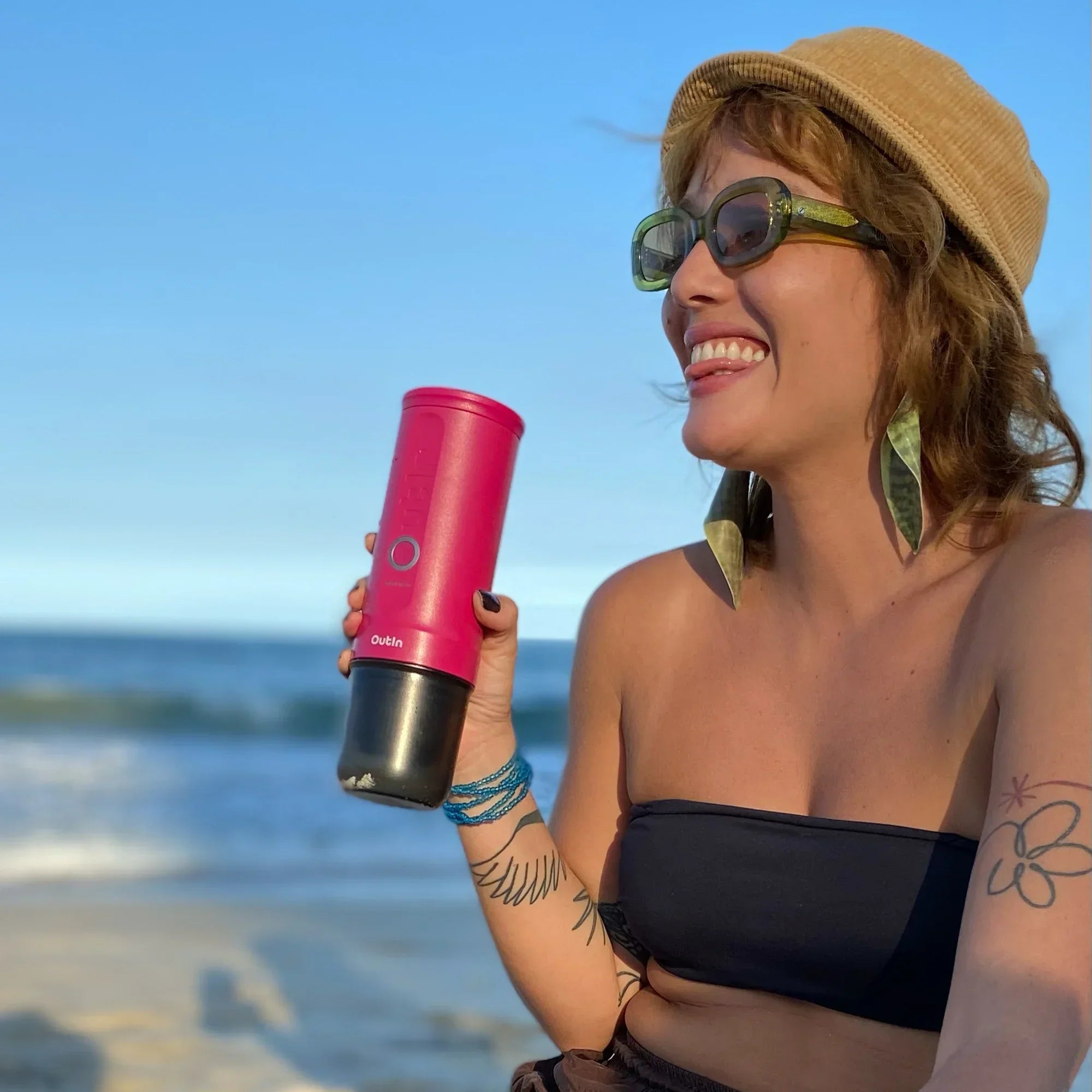 Woman enjoying beach with Fuchsia Pink Nano portable espresso machine, showcasing lifestyle portability.