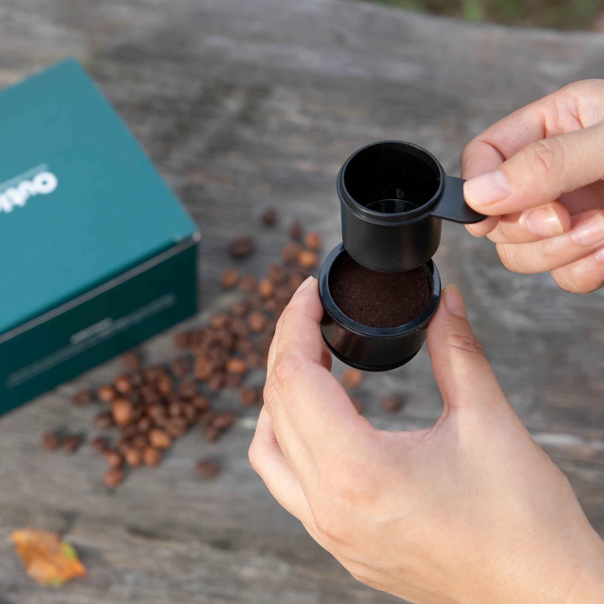Filter Basket and spoon over ground coffee, with a teal box beside.