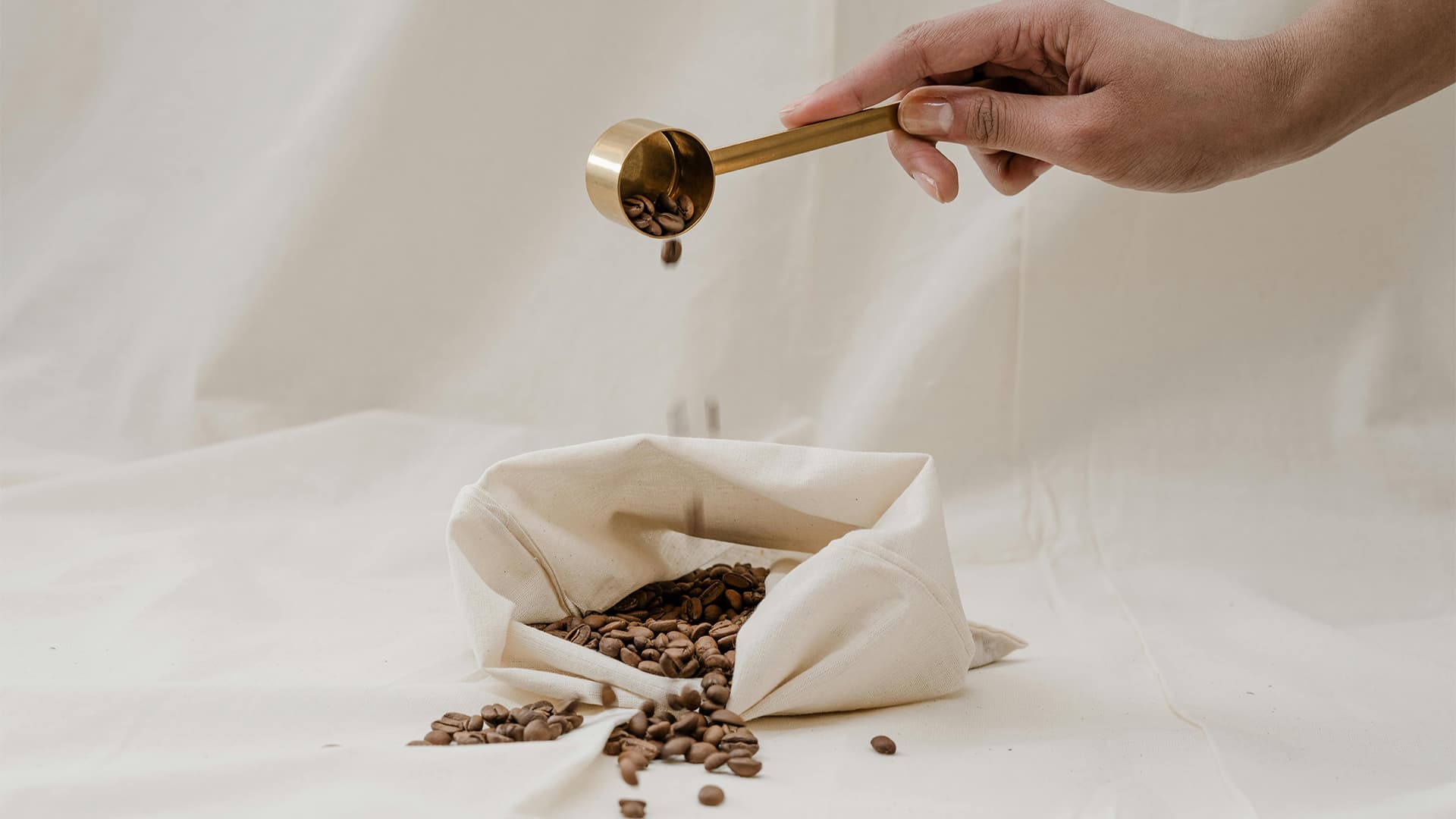 A hand uses a brass measuring scoop to drop whole coffee beans into a canvas storage bag on a neutral, textured surface