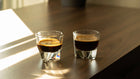 Two espresso shots in glass cups on a wooden table, bathed in warm sunlight with striped shadows