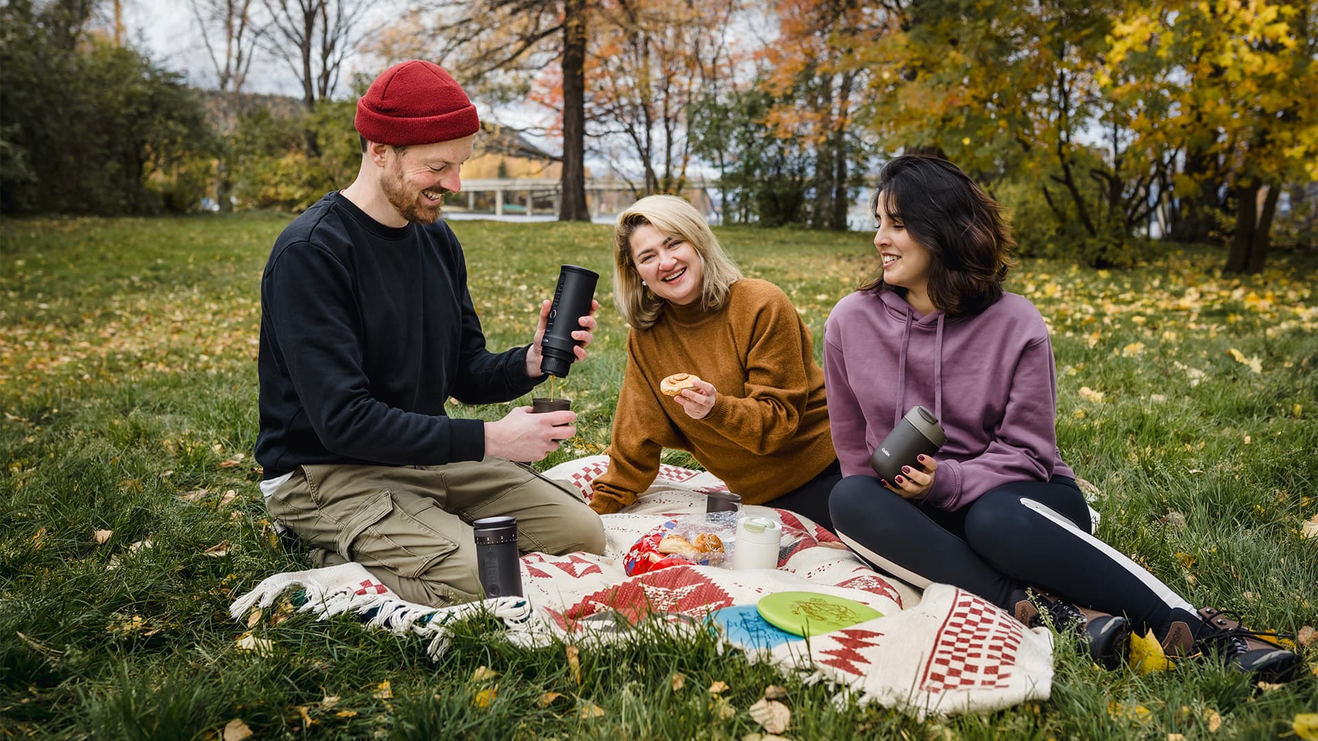 Group picnic in park, holding black portable coffee makers, enjoying autumn outdoors.