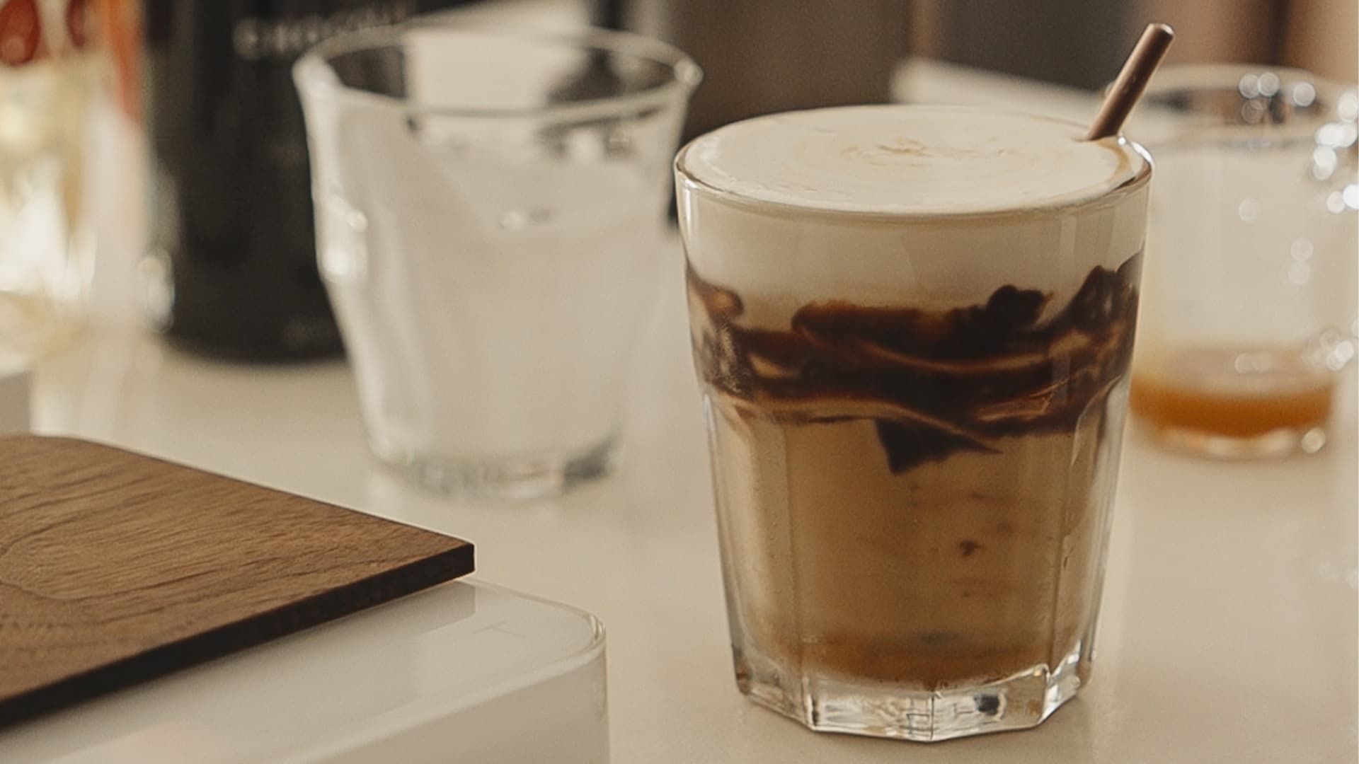 A close-up of an iced coffee drink in a faceted glass, topped with foam and chocolate drizzle, with a straw inserted