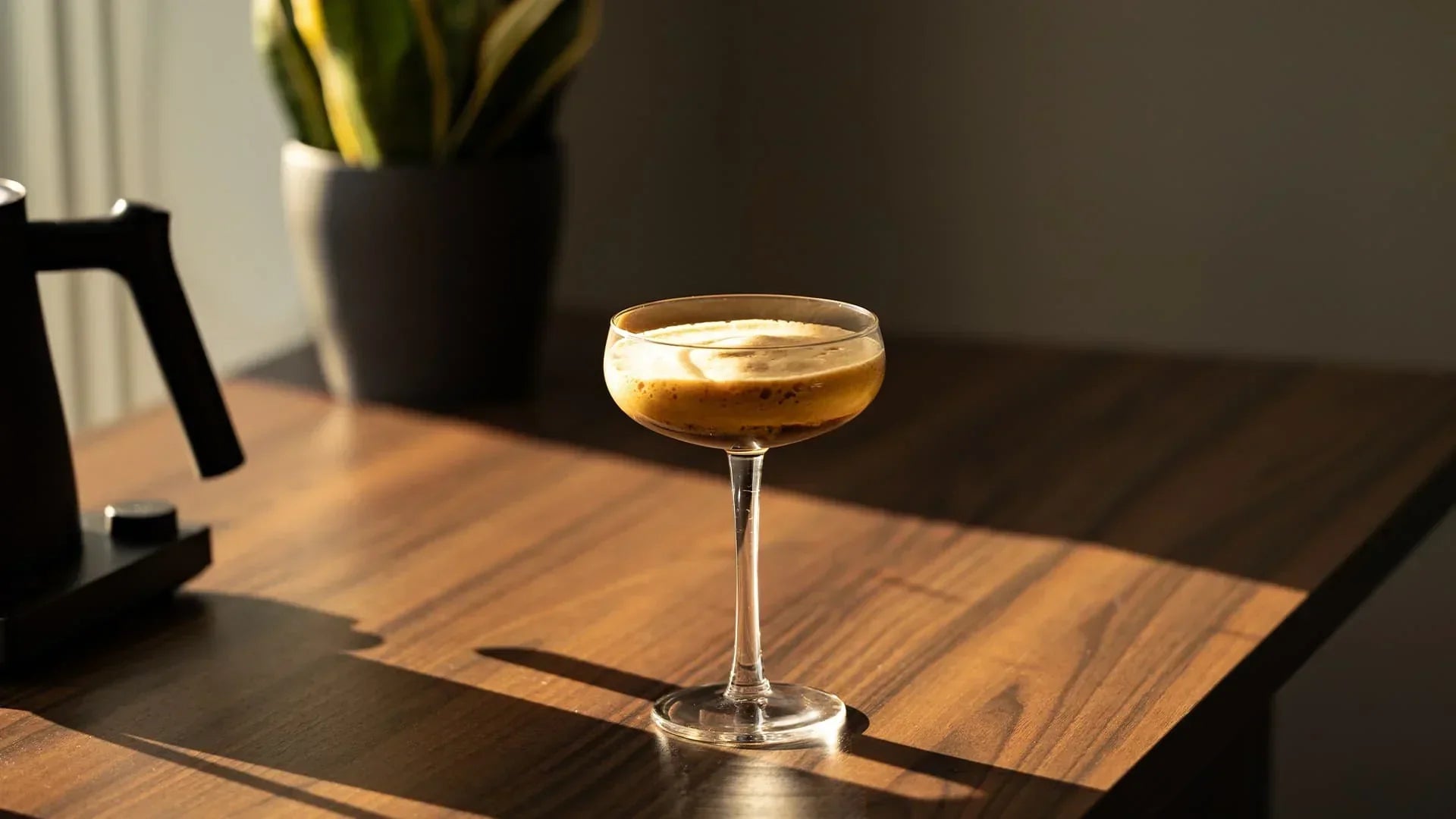 A foamy espresso drink in a stemmed glass sits on a sunlit wood table, casting long shadows. A potted plant is in the blurred background