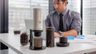An office worker uses the Outin Fino Portable Electric Coffee Grinder and other Outin accessories to brew coffee at his desk