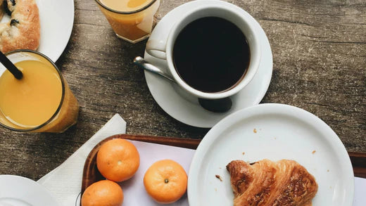 A breakfast scene with a cup of black coffee, fresh croissants and whole oranges on white plates atop a rustic wooden table.