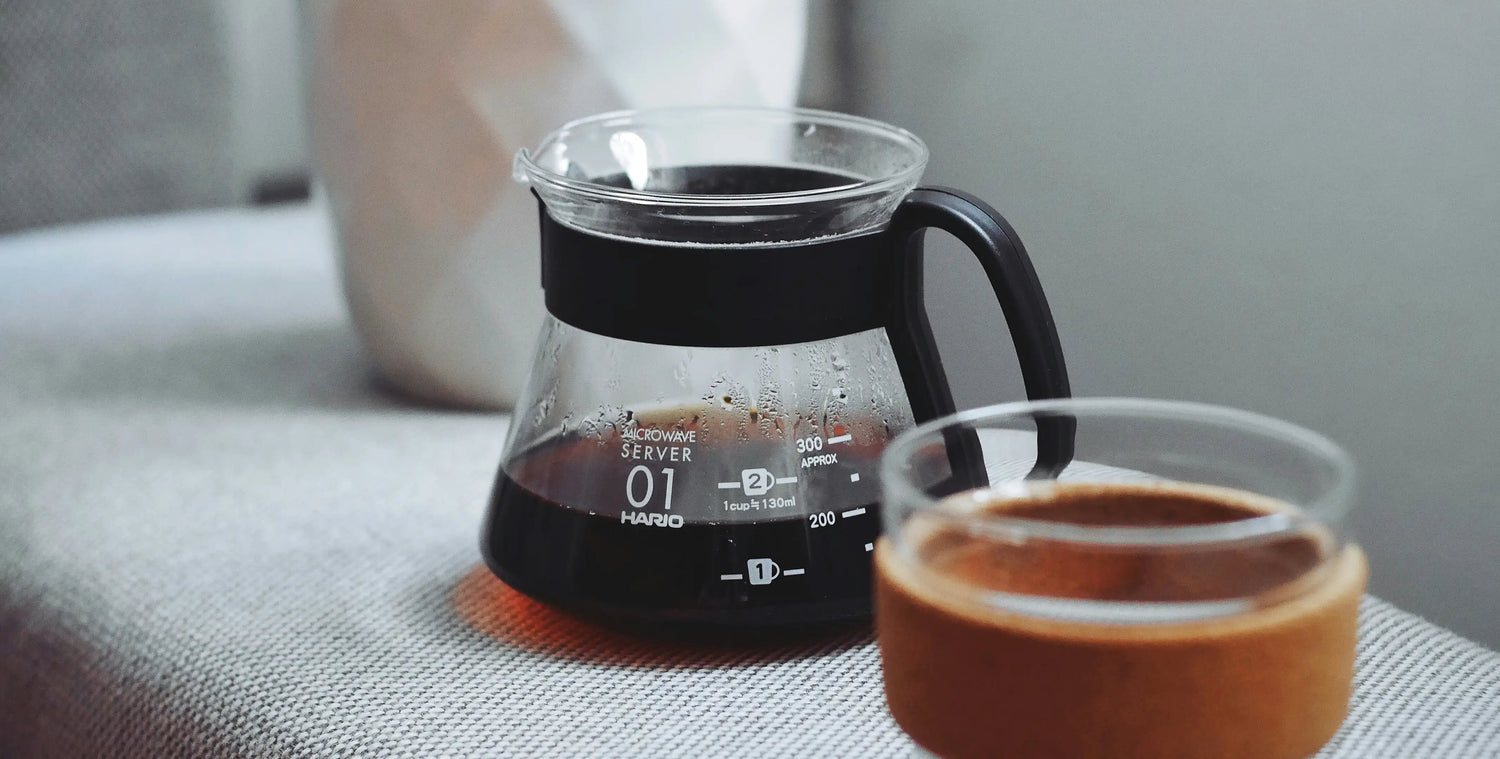 Glass coffee server with measurement markings and wooden lid next to a white mug on a textured surface