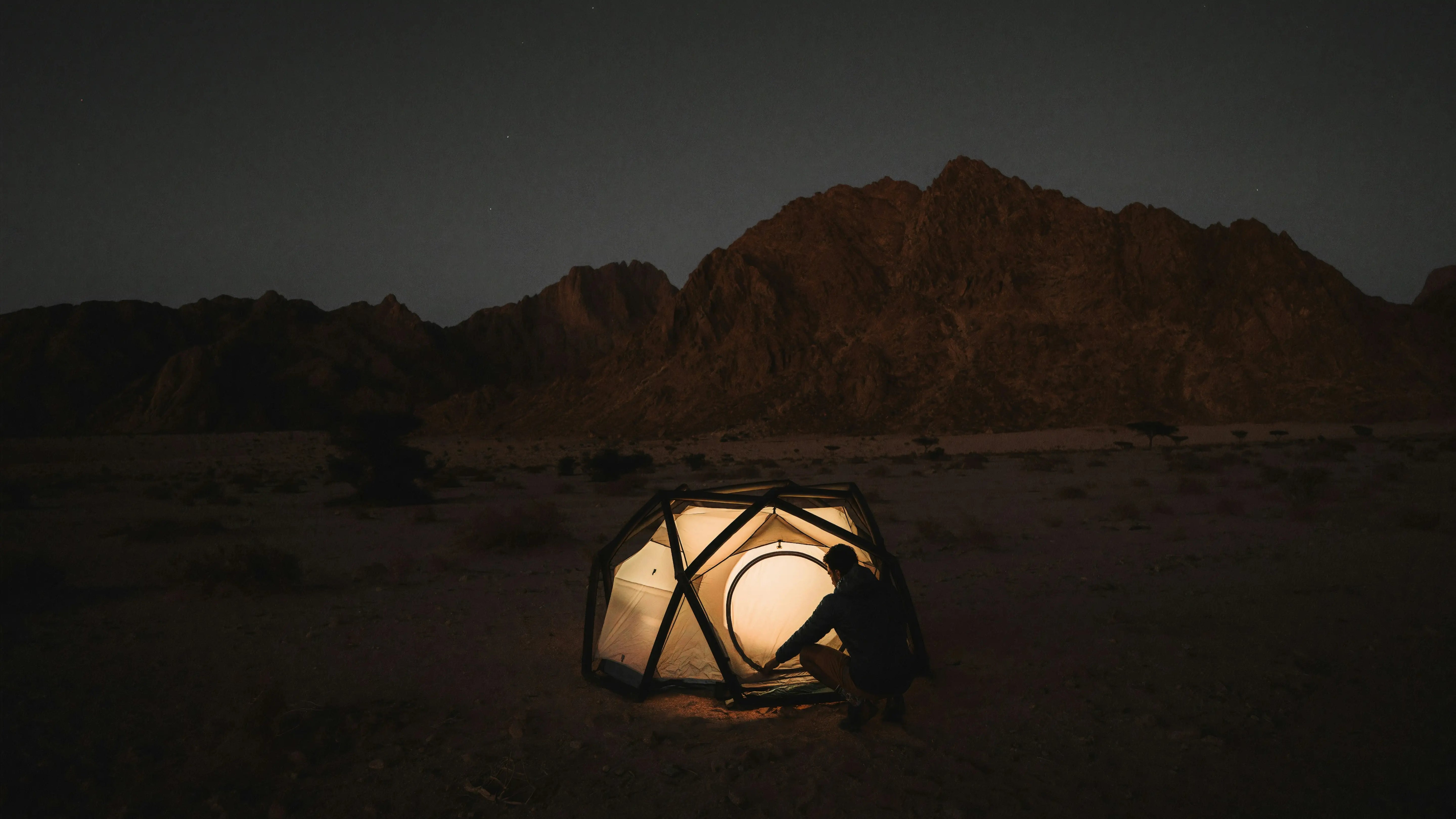 A glowing tent set up in a mountainous area at night, with a starry sky visible in the background.