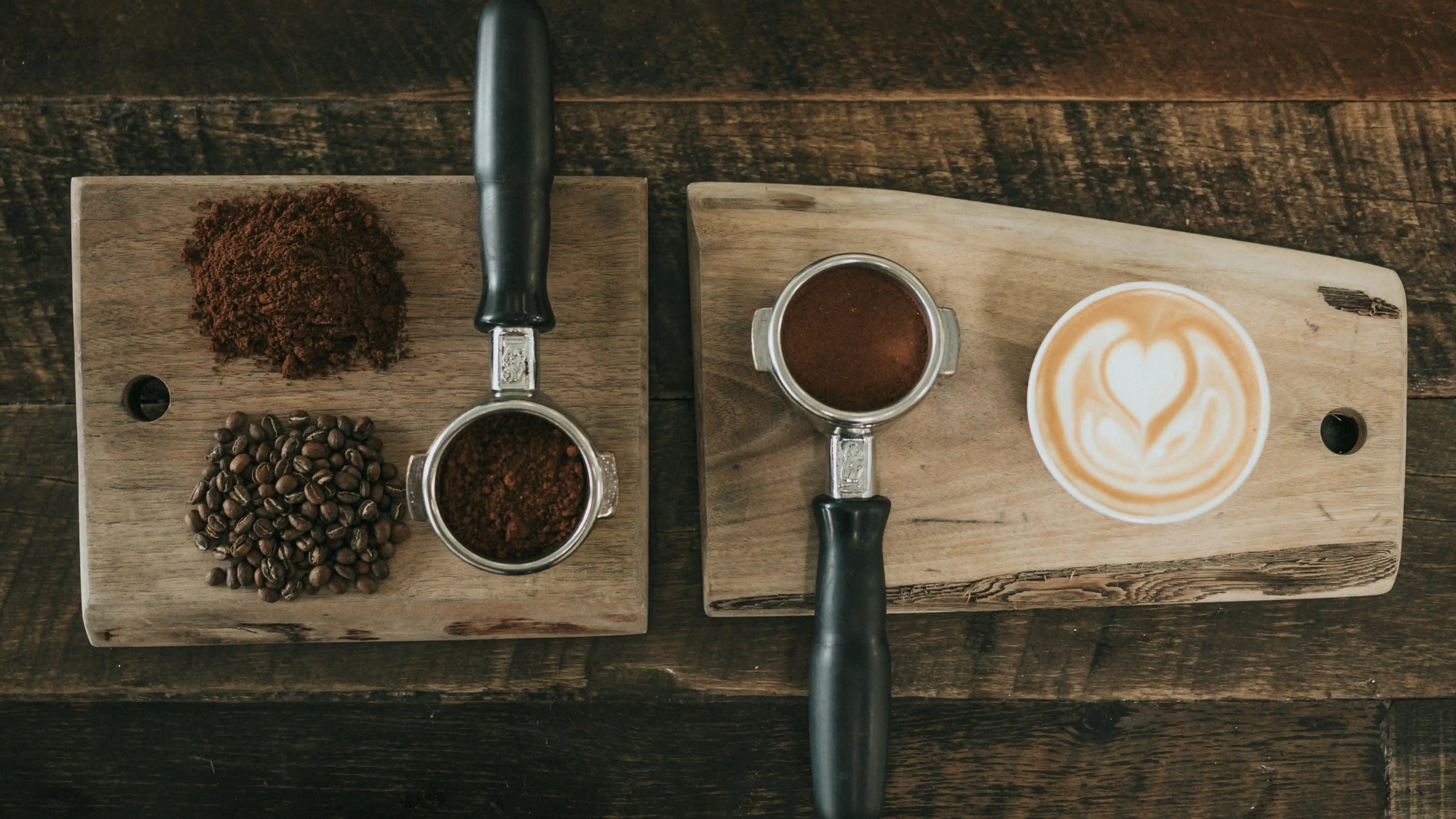 Overhead view of a coffee setup on a rustic wooden tray, featuring portafilters with ground coffee, coffee beans, and a latte art heart in a white cup.