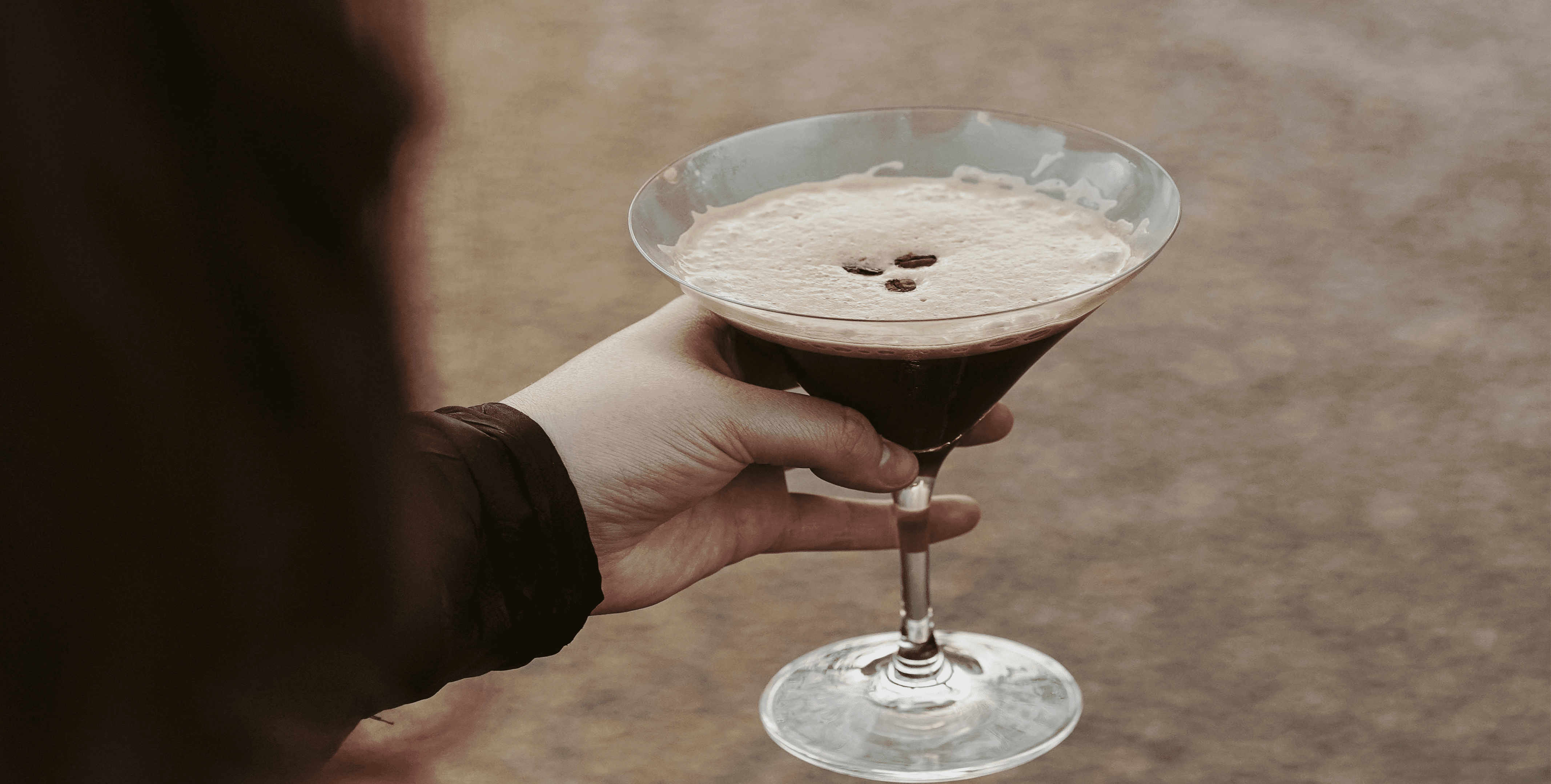 Hand holding an espresso martini in a wide-rimmed cocktail glass, topped with creamy foam and coffee beans, warm sepia tones