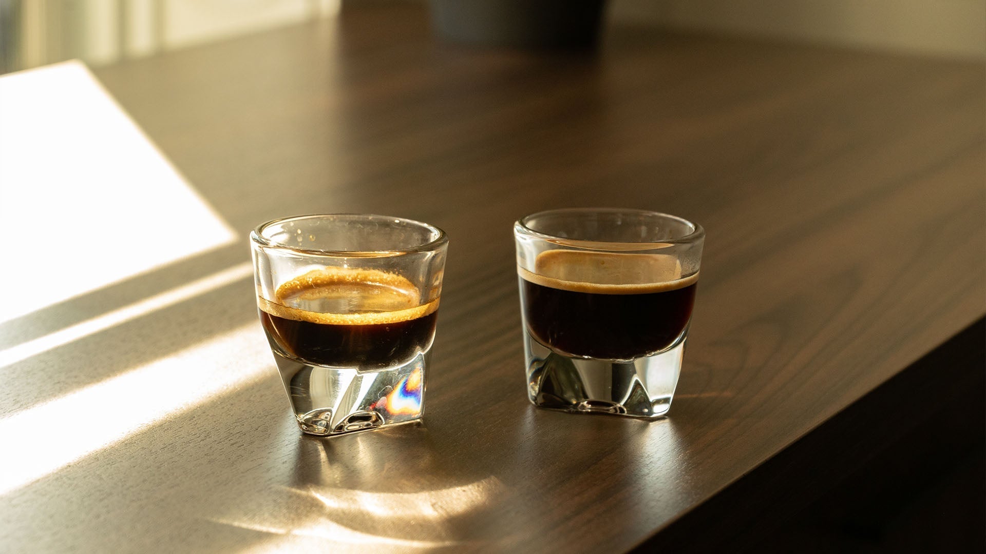 Two freshly pulled espresso shots in clear glass cups on wooden table bathed in warm morning sunlight. Golden crema visible on top shows perfect extraction, ideal base for summer coffee drinks.