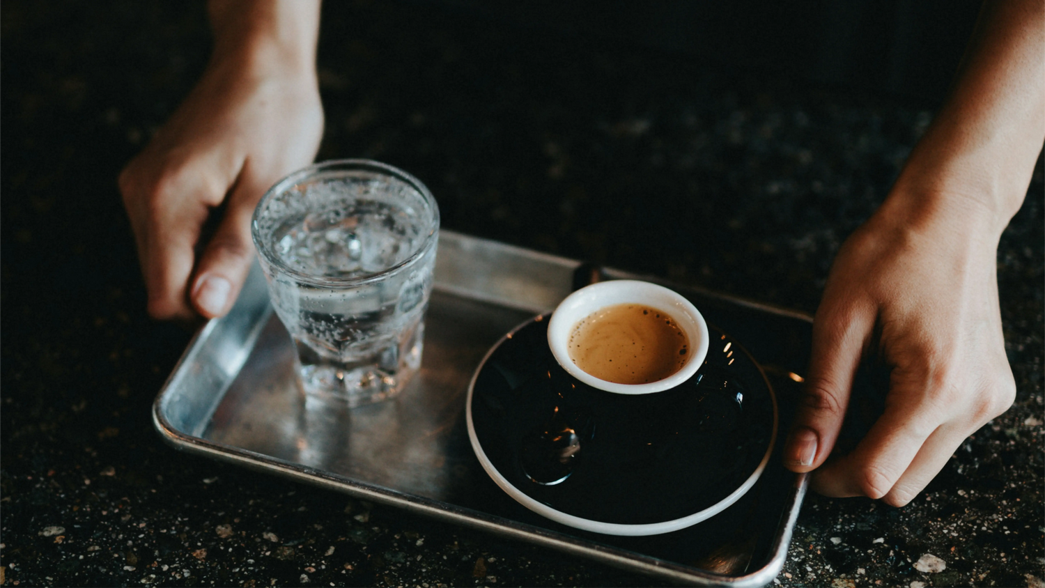 Perfect ristretto espresso served in black cup with sparkling water on metal tray, showing ideal 15-20ml concentrated shot volume