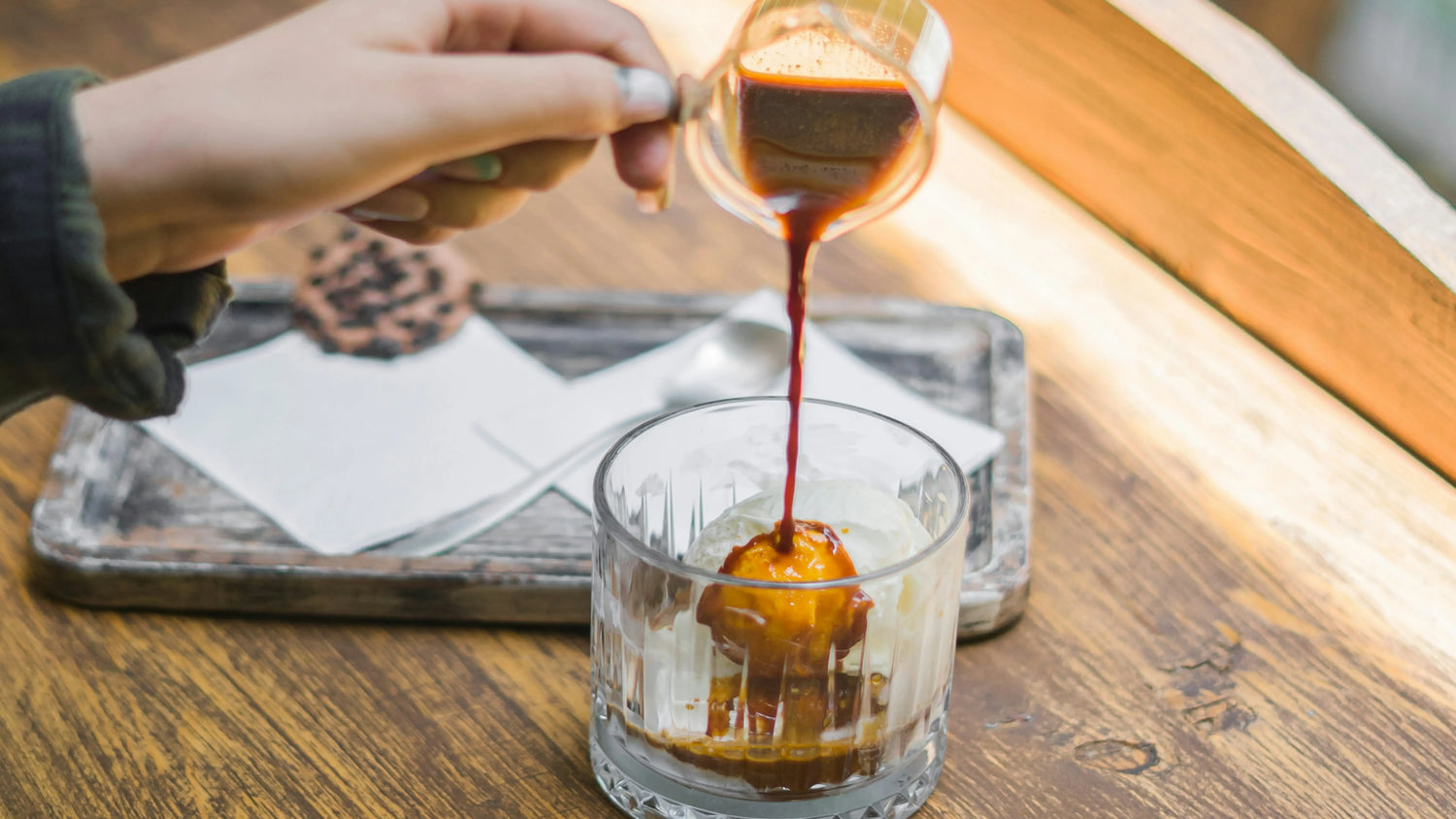 Hands preparing an iced espresso shot, pouring the fresh espresso from a glass into a rocks glass filled with ice on a wooden table.