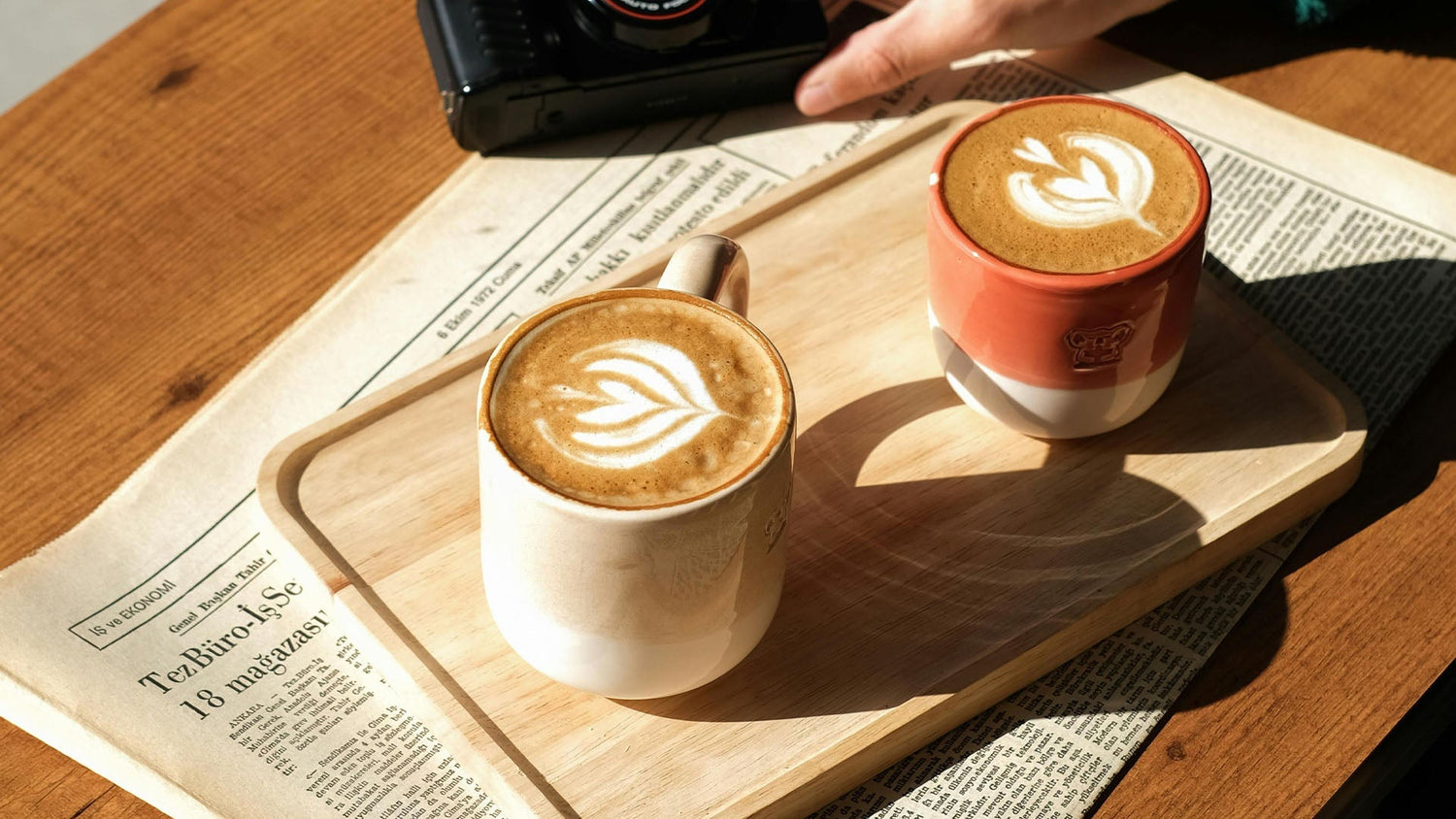 Two coffee cups with heart latte art on wooden serving board, natural sunlight streaming across newspaper background, creating warm cafe atmosphere