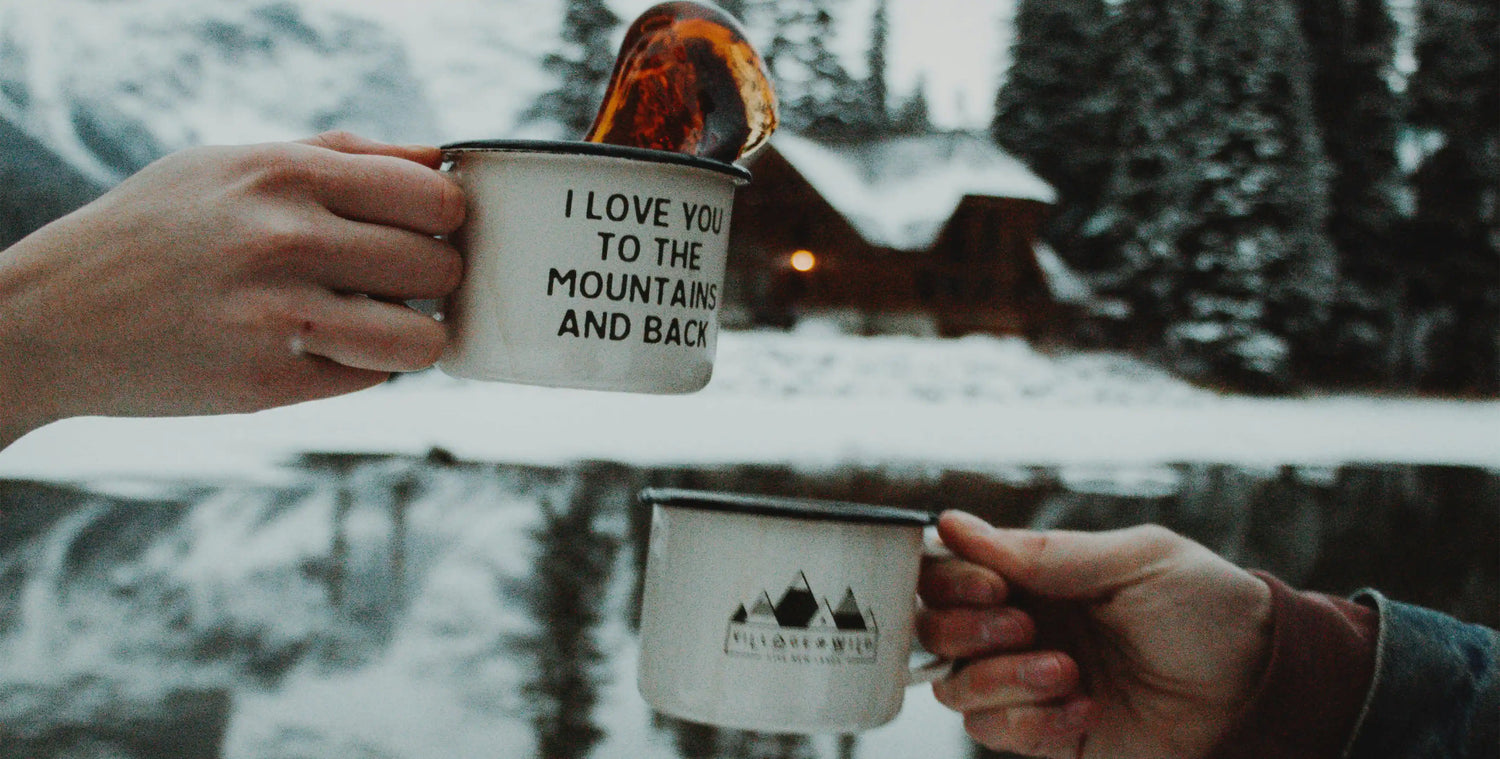 Two hands holding camping mugs with mountains in background