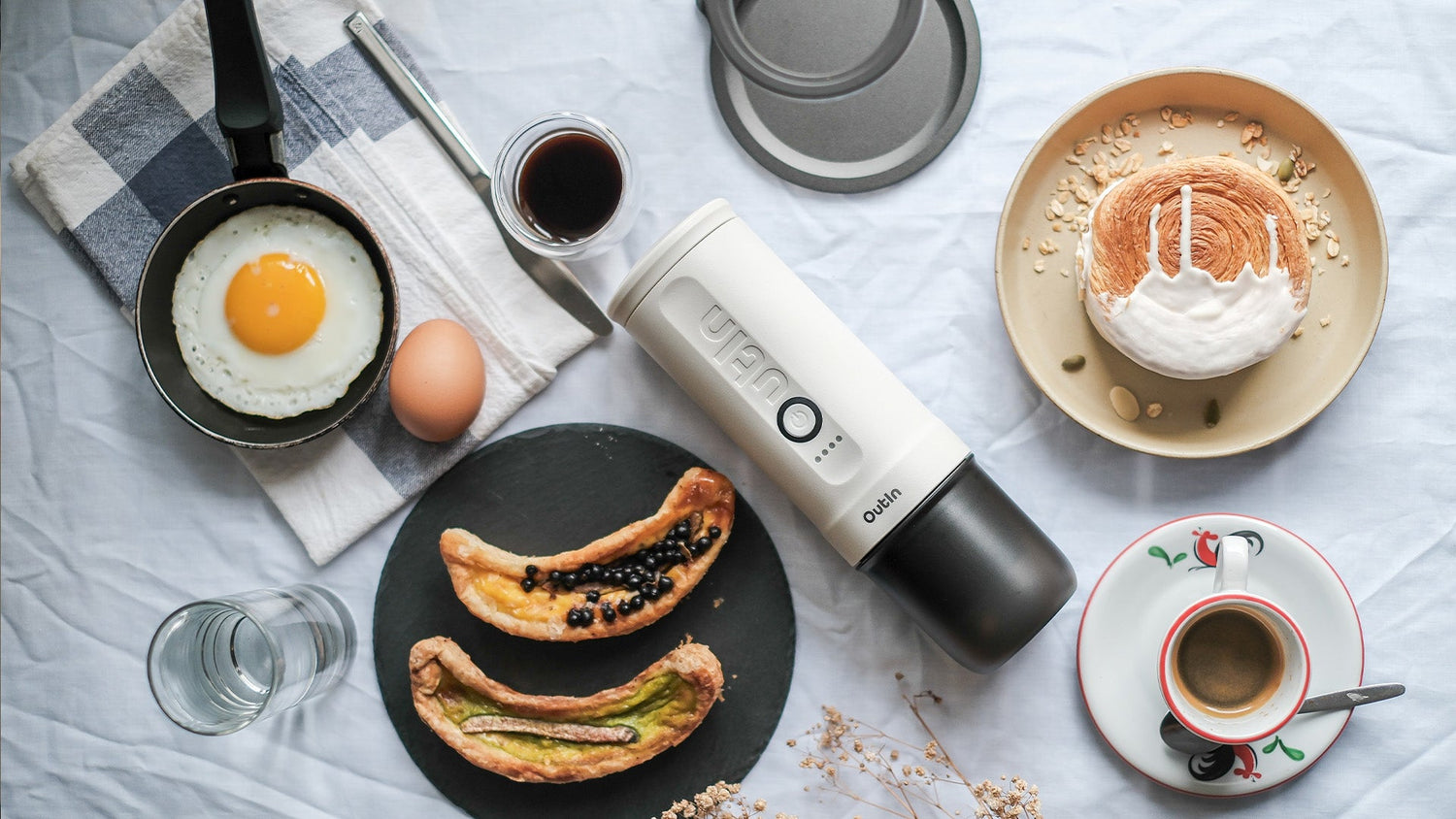 Breakfast scene with Outin portable espresso maker, fried egg, pastries and coffee cups on white tablecloth, illustrating how coffee consumption pairs with morning meals for optimal digestive benefits.