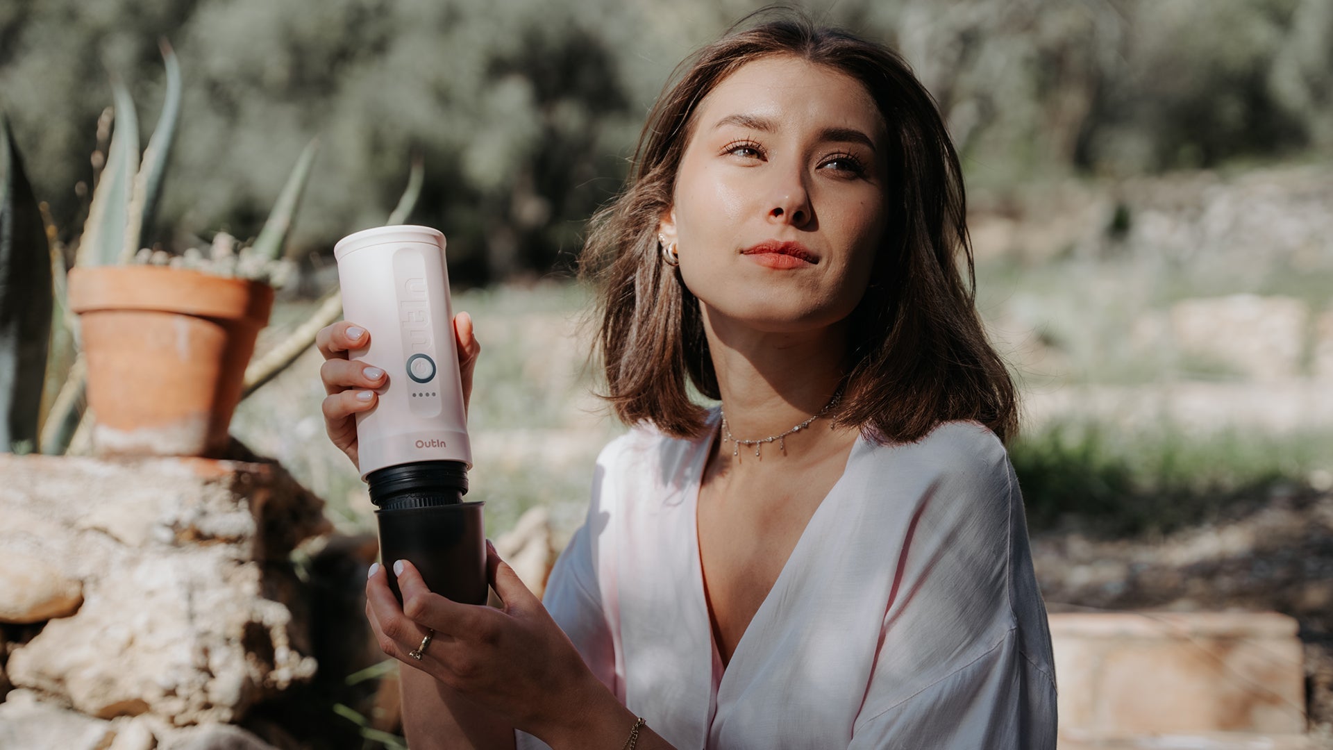 Beautiful woman in white outfit holding Outin Pink Edition Nano portable espresso maker outdoors, demonstrating its portability and stylish design while supporting breast cancer research for Women's Day.