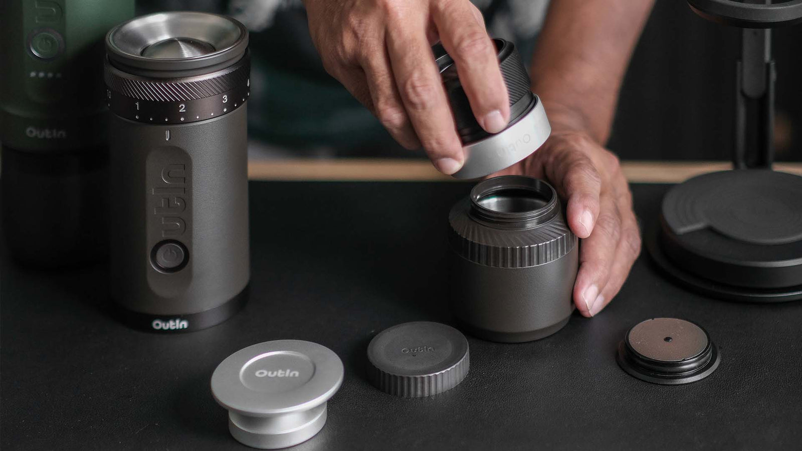 A person is shown assembling a dark gray Outin portable coffee grinder, holding the top grinding component and fitting it onto the main body of the device.