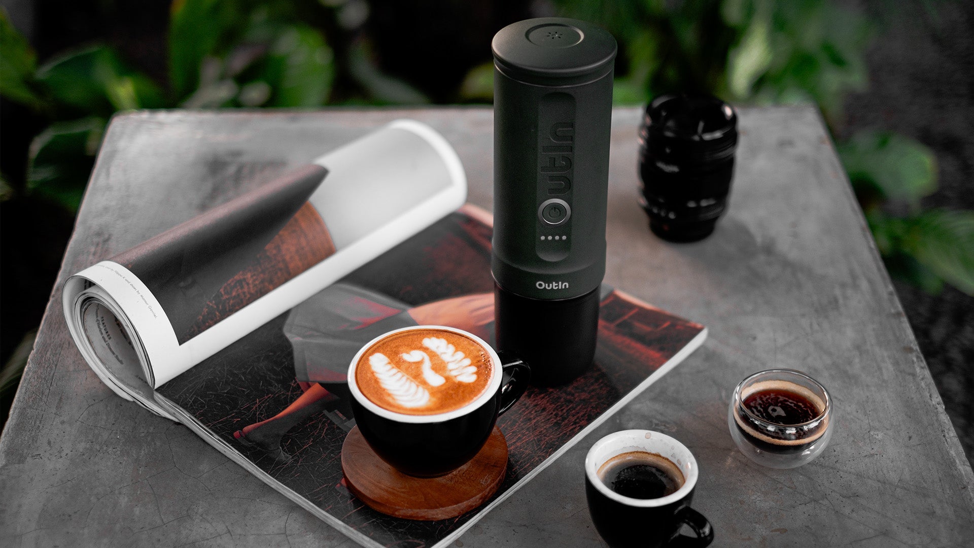 Outin portable espresso maker displayed with latte art coffee cup, magazine, and espresso shots on concrete table, showcasing strong coffee brewing options for caffeine enthusiasts.