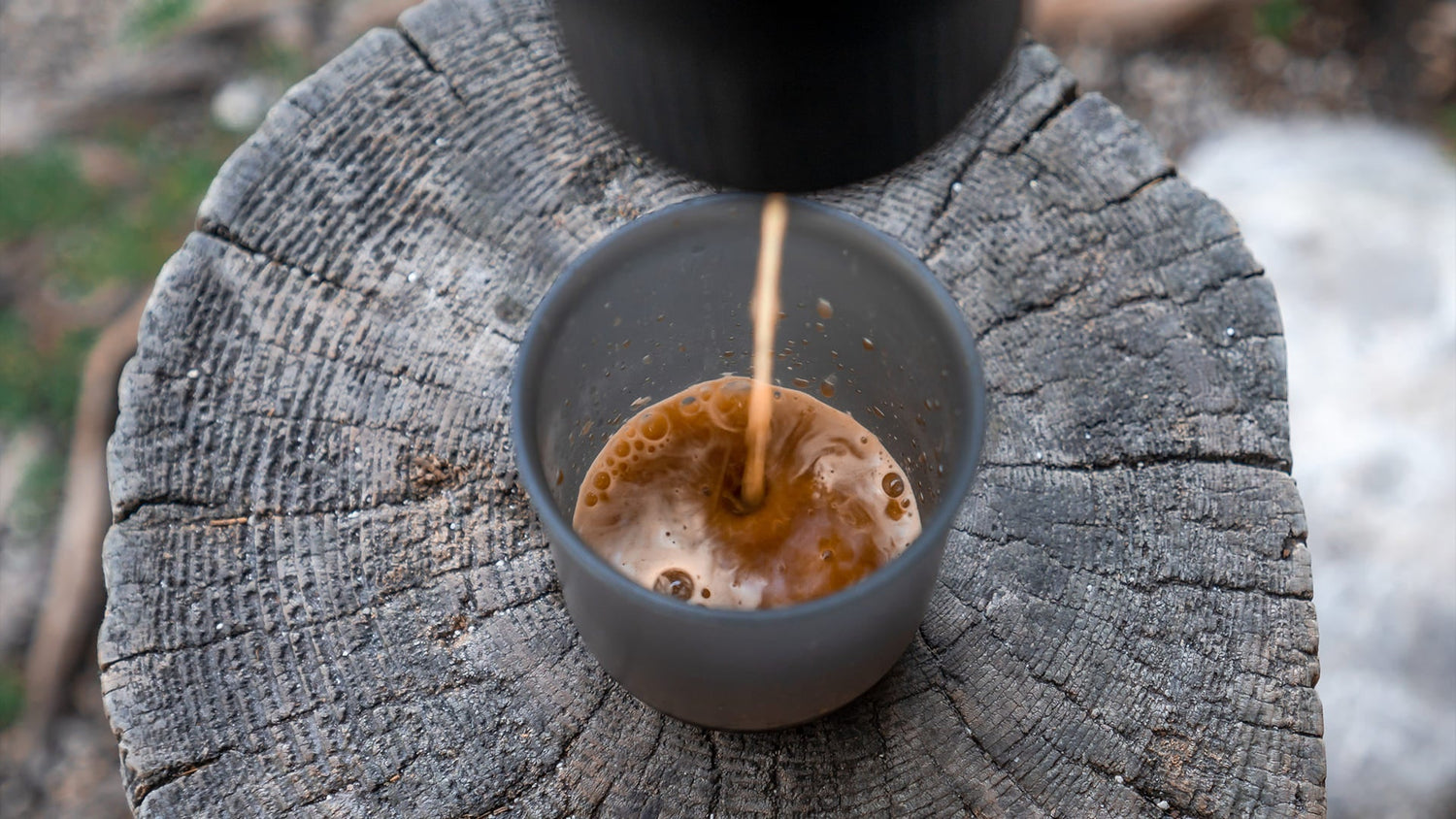 Fresh coffee being poured into a gray cup resting on a wooden stump, showing rich amber color and crema forming during the brewing process outdoors.