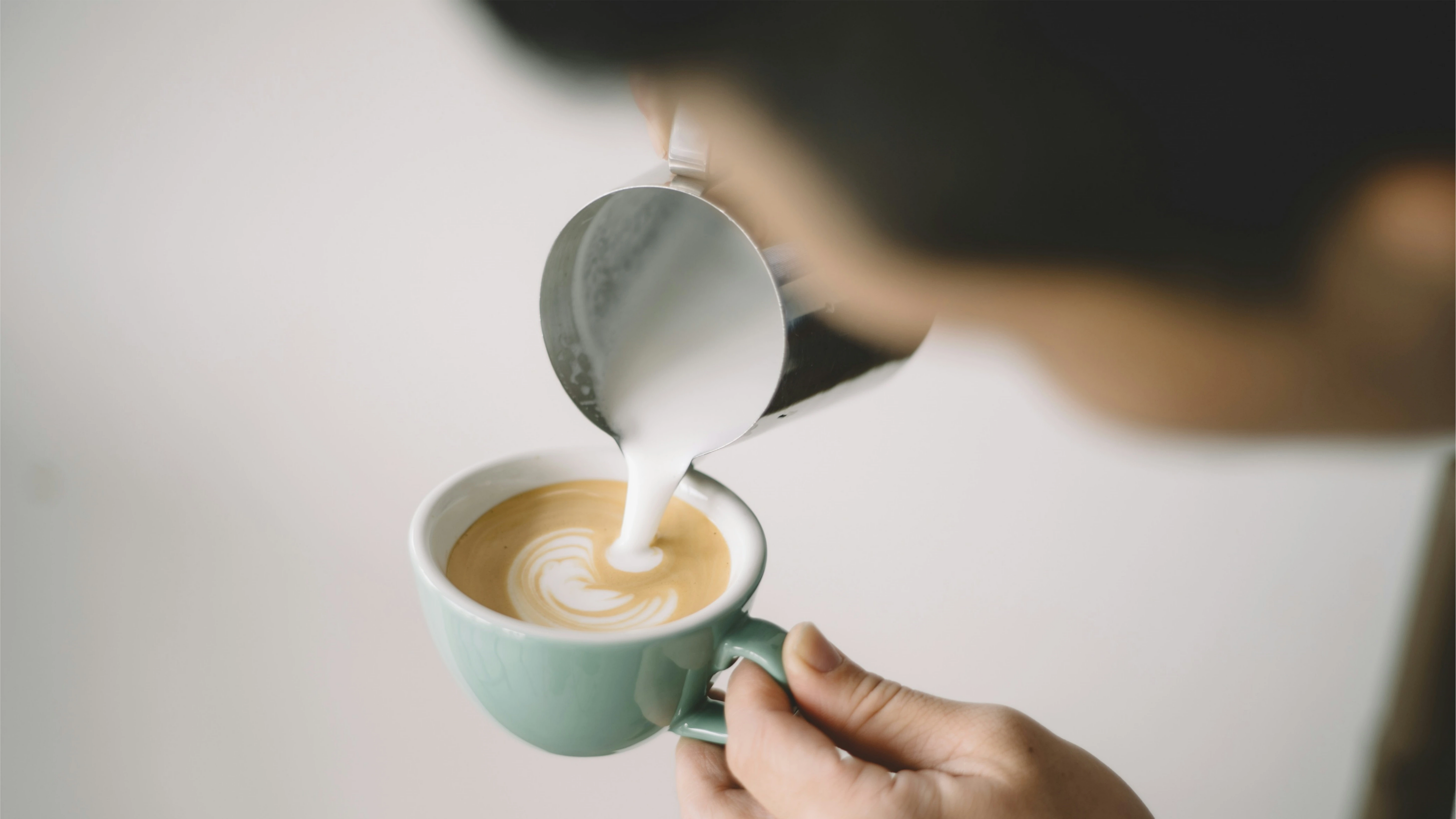 Pouring steamed low-fat milk into mint green cup creating latte art, demonstrating proper technique for making skinny latte with creamy microfoam texture