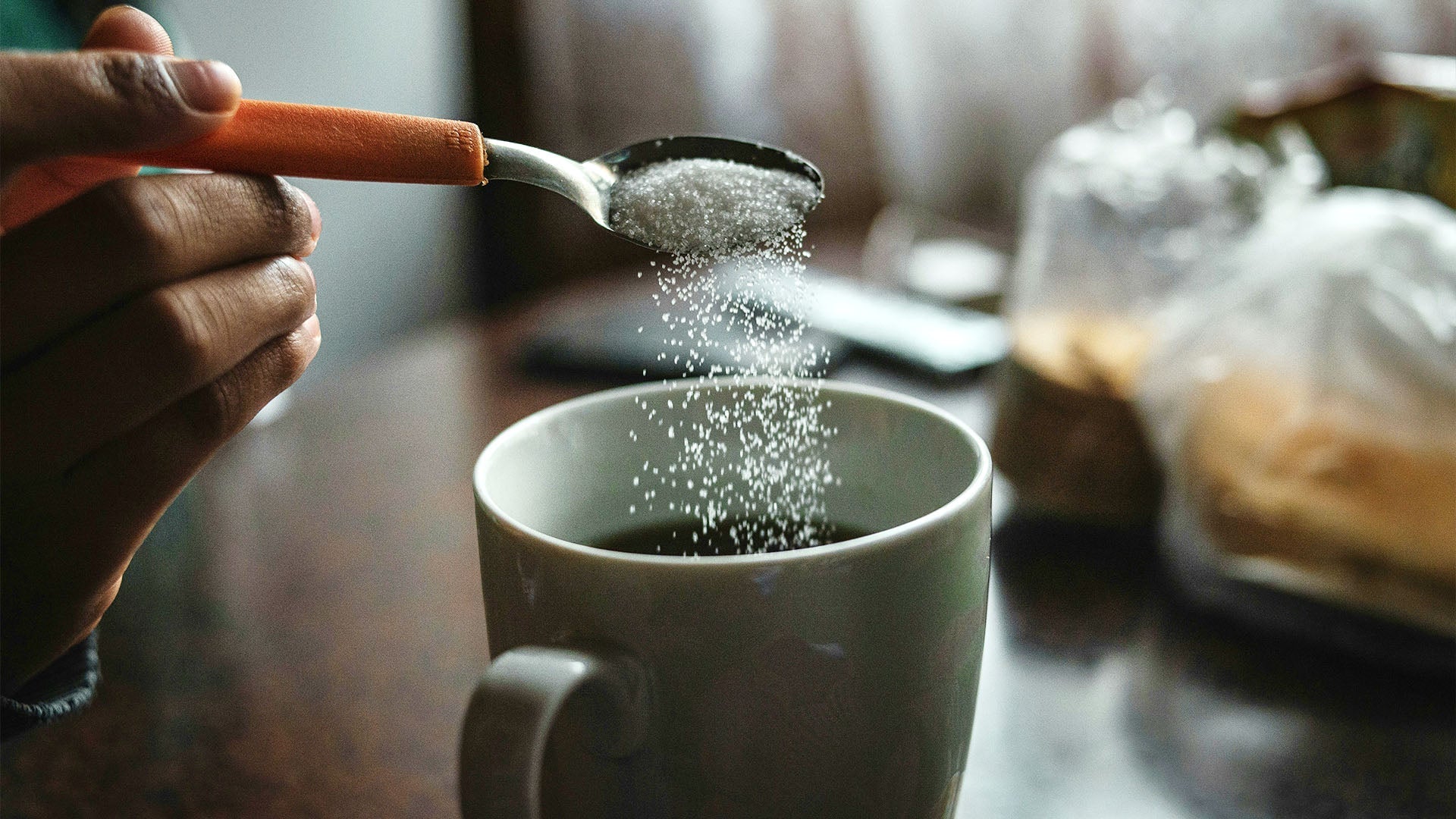 Hand sprinkling sugar into coffee cup, demonstrating traditional sweetening method. The article suggests using salt instead to reduce bitterness and enhance natural sweetness, allowing for less sugar consumption.