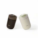 Two OutIn travel tumblers: one dark brown with a matching lid, and one cream with a contrasting light green lid.
