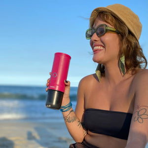 Smiling woman holding fuchsia Nano Portable Espresso Machine at the beach.